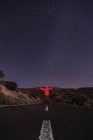 Viaggiatore in giacca rossa con cappuccio in piedi su strada vuota di notte con le braccia tese — Foto stock