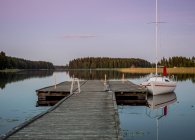 Pequeno iate branco com velas para baixo no cais de madeira à beira do lago perto de uma pequena aldeia na Finlândia — Fotografia de Stock