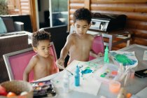 Deux garçons afro-américains torse nu utilisant une peinture brillante pour faire des images abstraites sur la table à la maison — Photo de stock