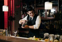 Jeune barman élégant travaillant derrière un comptoir de bar mélangeant des boissons dans un shaker — Photo de stock