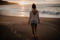 Вид ззаду на молоду жінку в повсякденному вбранні, що ходить на піщаному пляжі до штормового моря під час заходу сонця в природі — стокове фото