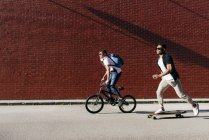 Side view of young black successful male friends in casual apparel and stylish sunglasses riding bike and skateboard — Stock Photo