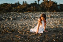 Bonito garoto feminino pensativo em vestido branco brincando com areia à beira-mar à luz do sol — Fotografia de Stock