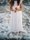 Portrait of crop charming little girl in white dress standing in water on beach — Stock Photo