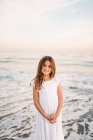 Portrait of charming little girl in white dress standing in water on beach and looking at camera — Stock Photo