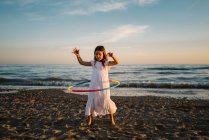 Little girl in white dress playing on seashore with hula hoop on background of evening sky — Stock Photo