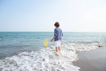 Боковой вид длинных волос симпатичный мальчик летом носить стоя в воде с рыболовной сеткой на берегу моря в солнечный день — стоковое фото
