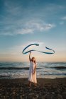 Little girl in white dress playing with long blue band on background of evening sky — Stock Photo