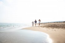 Niños felices y sonrientes en ropa casual corriendo descalzos a lo largo de la orilla del mar en la playa de arena en el día soleado de verano - foto de stock