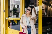 Молода чуттєва жінка в стильному вбранні і сонцезахисних окулярах стоїть біля кафе і дивиться вбік — стокове фото