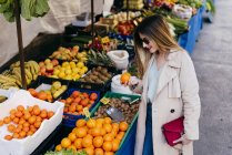 Молода щаслива красива жінка в стильному одязі і сонцезахисних окулярах, вибираючи фрукти на ринку на вулиці — стокове фото