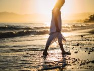 Маленька дівчинка в білій сукні, що йде на березі моря на фоні заходу сонця — стокове фото