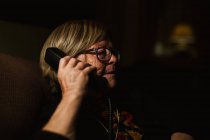 Feliz anciana sonriendo y respondiendo a la llamada telefónica mientras está sentada en la habitación oscura por la noche en casa - foto de stock