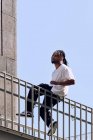 Молодой афроамериканец сидит на перилах моста на городской улице в солнечный день — стоковое фото