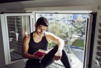 Біля вікна сидить молодий поважний чоловік у звичайній безрукавній сорочці і читає цікаву книжку. — стокове фото