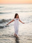 Niña en vestido blanco caminando y jugando en la orilla del mar en el fondo de la puesta del sol - foto de stock