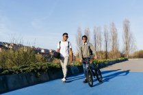 Молодой афроамериканец улыбается успешным мужчинам в повседневной одежде и стильных солнцезащитных очках, прогуливаясь по городу на велосипеде и скейтборде — стоковое фото