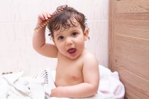Очаровательный ребенок смотрит в камеру и расчесывает влажные волосы, сидя на полотенце в ванной комнате после душа — стоковое фото