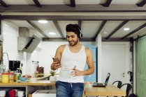 Young cheerful successful man in headphones leaning on chair, browsing smartphone and holding cup of hot coffee in kitchen — Stock Photo
