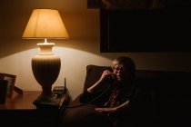 Heureuse femme âgée souriant et répondant à un appel téléphonique alors qu'elle était assise dans une pièce sombre le soir à la maison — Photo de stock