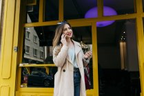 Молодая веселая женщина, стоящая возле кафе и разговаривающая по смартфону — стоковое фото