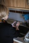 Visão traseira da mulher idosa em pé perto da pia e lavagem de vidro com sabão na cozinha em casa — Fotografia de Stock