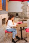 Jovem do sexo feminino em roupa casual sentado à mesa no café ao ar livre e smartphone de navegação na rua da cidade — Fotografia de Stock