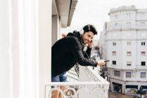 Giovane sorridente bel maschio casual in auricolare appoggiato su ringhiera terrazza con smartphone e guardando altrove — Foto stock