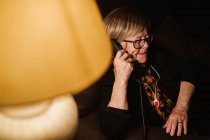 Glad elderly woman smiling and answering phone call while sitting in dark room in evening at home — Stock Photo