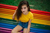 Jolie jeune femme en tenue décontractée souriant joyeusement et regardant la caméra tout en étant assis sur le banc arc-en-ciel dans le parc — Photo de stock