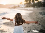 Joyeux enfant féminin mignon en robe blanche s'amusant sur le bord de mer sablonneux au coucher du soleil — Photo de stock