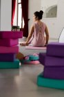 Vista posteriore di bruna sottile in abbigliamento sportivo rosa seduto sul tappeto accanto a attrezzature colorate per lo yoga al chiuso — Foto stock