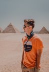 Fiducioso viaggiatore maschile in occhiali da sole guardando lontano mentre in piedi nel deserto contro le famose Grandi Piramidi al Cairo, Egitto — Foto stock