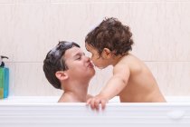 Вид сбоку мальчика, надувающего пузырь на лице симпатичного ребенка во время совместного купания — стоковое фото