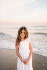 Portrait of charming little girl in white dress standing on sandy beach and looking at camera — Stock Photo