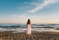 Back view of cute female child in white dress standing on sandy seashore — Stock Photo