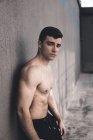 Мускулистый парень без рубашки смотрит в камеру, опираясь на цементную стену во время тренировки на городской улице — стоковое фото