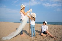 Chicas con sombreros adjuntando toldo en el poste, mientras que el niño sentado en la arena en la playa - foto de stock