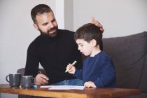 Adult bearded man helping little boy to draw picture while sitting at table at home together — Stock Photo