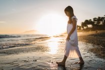 Вид сбоку маленькой девочки в белом платье, идущей по берегу моря на фоне солнца — стоковое фото