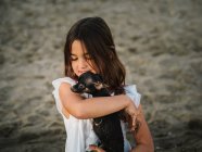 Retrato de una encantadora niña en vestido blanco sosteniendo a un perrito en la playa - foto de stock