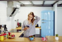 Junge verführerisch charmante Frau mit blonden Haaren in lässigem Hemd, die in der Küche steht und das Smartphone benutzt — Stockfoto