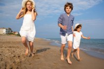 Счастливые и улыбающиеся дети в повседневной одежде бегают босиком по берегу моря на песчаном пляже в летний солнечный день — стоковое фото