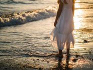 Маленька дівчинка в білій сукні стоїть на березі моря на фоні сонця — стокове фото