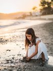 Lindo niño femenino pensativo en vestido blanco jugando con arena en la playa a la luz del sol - foto de stock