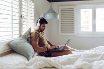 Молодий чоловік сконцентрований на успішній людині у звичайній сорочці та навушниках, сидячи на ліжку за допомогою ноутбука. — стокове фото