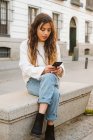 Junge Frau in lässigem Outfit sitzt und surft Smartphone auf der Straße der Stadt — Stockfoto