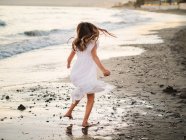 Little girl in white dress playing on seashore at sunset — Stock Photo