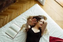 Jovem sorrindo mulher deitada na cama com livro e gesticulando com a mão — Fotografia de Stock