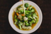 From above hand holding tasty broccoli with chopsticks over soup with carrot, onion and bell pepper in Asian restaurant — Stock Photo